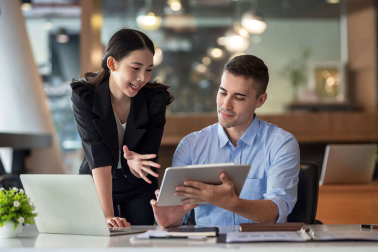 coworkers-discuss-work-project-over-tablet-and-laptop-at-desk-scaled